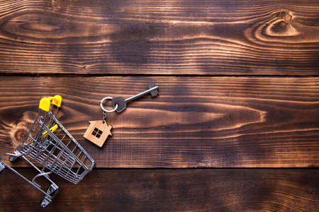 A key with a key ring in the shape of a house on a wooden background sticks out of a reduced supermarket grocery cart. Buying, renting a house, turnkey business, mortgage. Space for text. Flat layの写真素材