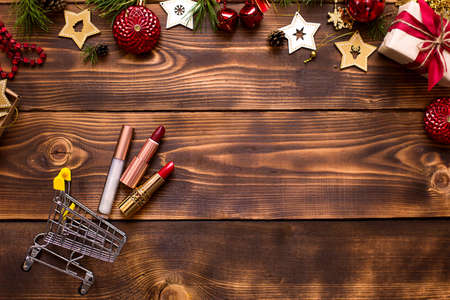Red lipstick and lip gloss and eyelids on a wooden background in metal supermarket cart in the Christmas decor. Holiday makeup for new year, gift, shopping, women's desires. Space for text, flat layの写真素材