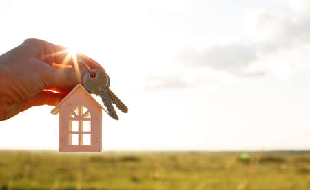 White wooden figure of a house and keys in hand against the background of the sky and field. Dream of your home, building a cottage in the countryside, plan and design, delivery of the project, moving to a new house.の写真素材