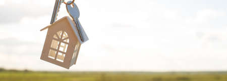 White wooden figure of a house and keys in hand against the background of the sky and field. Dream of your home, building a cottage in the countryside, plan and design, delivery of the project, moving to a new house.の写真素材