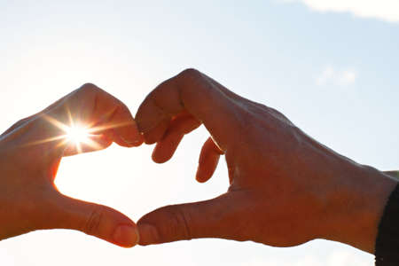 Two hands, male and female, are joined in the shape of a heart against the sky with the sun's rays at the opening. Symbol of a couple in love, Valentine's day, romance, wedding in heaven. copyspaceの写真素材