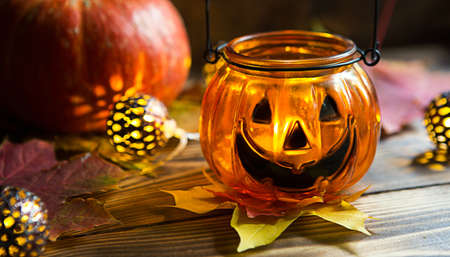 Lamp pumpkin with eyes and mouth made of glass and natural orange pumpkin on a wooden table with yellow and red maple leaves. Halloween, warm autumn atmosphere. Round garland, candle in a candlestick.の写真素材