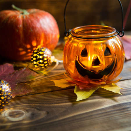 Lamp pumpkin with eyes and mouth made of glass and natural orange pumpkin on a wooden table with yellow and red maple leaves. Halloween, warm autumn atmosphere. Round garland, candle in a candlestick.の写真素材
