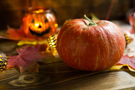 Lamp pumpkin with eyes and mouth made of glass and natural orange pumpkin on a wooden table with yellow and red maple leaves. Halloween, warm autumn atmosphere. Round garland, candle in a candlestick.の写真素材