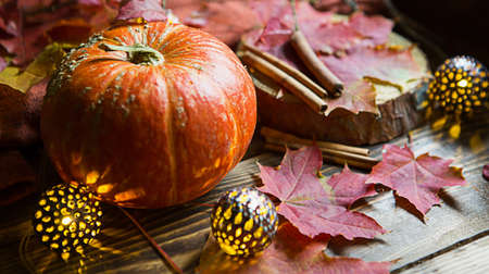 Orange natural round pumpkin on a wooden table with fallen yellow and red maple leaves, cinnamon sticks. Lights garlands, warm autumn atmosphere, thanksgiving, harvest festival, Halloween. Copy spaceの写真素材