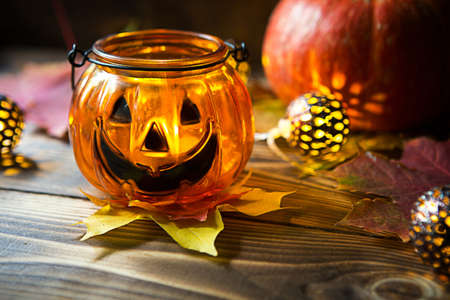 Lamp pumpkin with eyes and mouth made of glass and natural orange pumpkin on a wooden table with yellow and red maple leaves. Halloween, warm autumn atmosphere. Round garland, candle in a candlestick.の写真素材