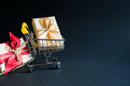 Box with a gift in Christmas packaging in a shopping cart on a black background. Black Friday, buying gifts for the new year. Space for textの写真素材