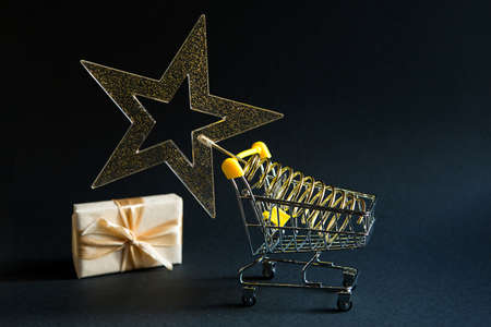 Shopping cart with a gold transparent star with sequins in the shape of a Christmas tree on a black background. Black Friday, buying gifts for the new year. Space for textの写真素材