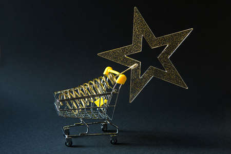Shopping cart with a gold transparent star with sequins in the shape of a Christmas tree on a black background. Black Friday, buying gifts for the new year. Space for textの写真素材
