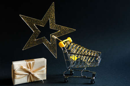 Shopping cart with a gold transparent star with sequins in the shape of a Christmas tree on a black background. Black Friday, buying gifts for the new year. Space for textの写真素材