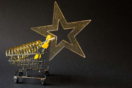 Shopping cart with a gold transparent star with sequins in the shape of a Christmas tree on a black background. Black Friday, buying gifts for the new year. Space for textの写真素材
