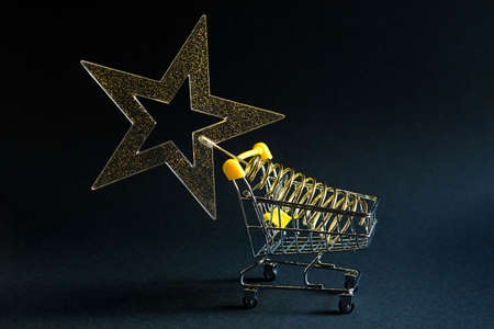 Shopping cart with a gold transparent star with sequins in the shape of a Christmas tree on a black background. Black Friday, buying gifts for the new year. Space for textの写真素材