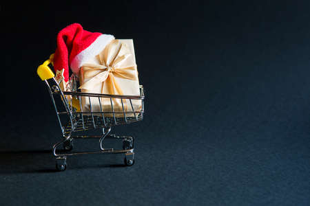 Box with a gift in Christmas packaging in a shopping cart on a black background. Black Friday, buying gifts for the new year. Space for textの写真素材
