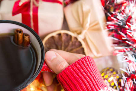 A woman's hand in a warm sweater holds a red mug with a hot drink on a table with Christmas decorations. New year's atmosphere, cinnamon sticks and a slice of dried orange, gifts, garland and tinselの写真素材