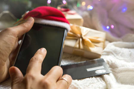 Women's hands with smartphone in Santa hat, Bank card on the background of Christmas decorations, gift boxes, garlands. New year's shopping, buying gifts through the app, online store. Online shoppingの写真素材
