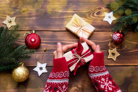 Women's hands in knitted sweater with ornament hold a box with Christmas gift in their palms on the table among the decor. New year, packaging and preparation of gifts, branches of fir, decorationの写真素材