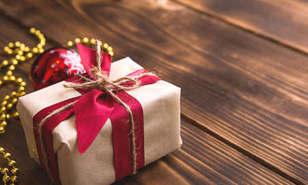 A box with a Christmas gift in light paper is tied with a red ribbon on a wooden background with decor. Christmas tree pendant, beads, garland. New Year. space for textの写真素材