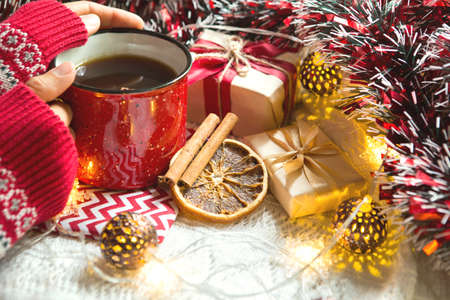 A woman's hand in a warm sweater holds a red mug with a hot drink on a table with Christmas decorations. New year's atmosphere, cinnamon sticks and a slice of dried orange, gifts, garland and tinselの写真素材