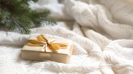A box with a Christmas gift in light paper is tied with a gold ribbon on a white knitted background with decor. Christmas tree pendant, green branches, lights garlands. New Year. space for textの写真素材