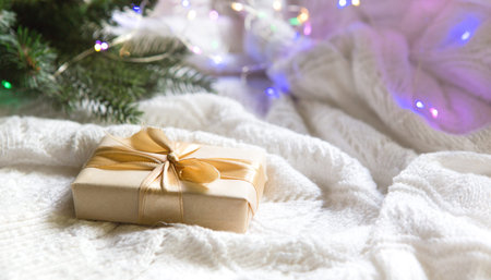A box with a Christmas gift in light paper is tied with a gold ribbon on a white knitted background...