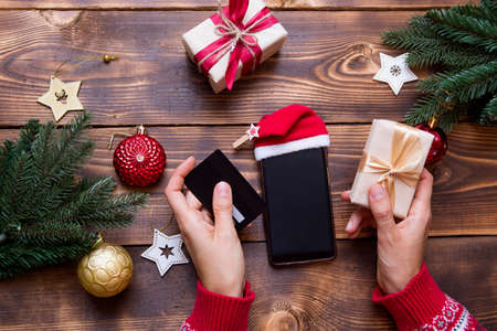 Women's hands with smartphone in Santa hat, Bank card on the background of Christmas decorations, gift boxes, garlands. New year's shopping, buying gifts through the app, online store. Online shoppingの写真素材