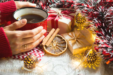 A woman's hand in a warm sweater holds a red mug with a hot drink on a table with Christmas decorations. New year's atmosphere, cinnamon sticks and a slice of dried orange, gifts, garland and tinselの写真素材