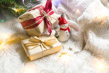Boxes with Christmas gifts and decorations snowman on a white knitted blanket in the lights of garlands and spruce branches. New year, festive atmosphere. Space for textの写真素材