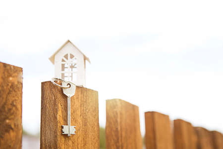 Small figure of white wooden house on fence with key to housing against the background of cottages. Building, design, project, moving to new house, mortgage, rent and purchase real estate. Copy spaceの写真素材