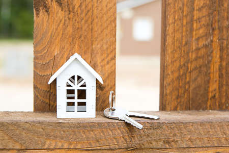 Small figure of white wooden house on fence with key to housing against the background of cottages. Building, design, project, moving to new house, mortgage, rent and purchase real estate. Copy spaceの写真素材