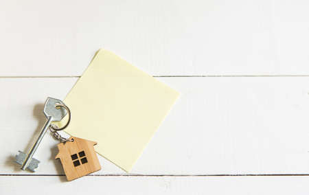 Keychain in the shape of wooden house with key on a white background with a square sheet for notes. Building, design, project, moving to new house, mortgage, rent and purchase real estate. Copy spaceの写真素材