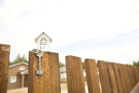 Small figure of white wooden house on fence with key to housing against the background of cottages. Building, design, project, moving to new house, mortgage, rent and purchase real estate. Copy spaceの写真素材