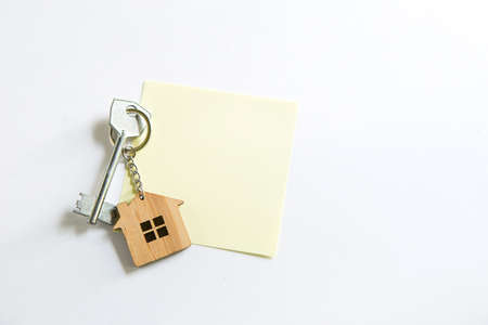 Keychain in the shape of wooden house with key on a white background with a square sheet for notes. Building, design, project, moving to new house, mortgage, rent and purchase real estate. Copy spaceの写真素材