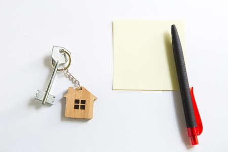 Keychain in the shape of wooden house with key on a white background with a square sheet for notes. Building, design, project, moving to new house, mortgage, rent and purchase real estate. Copy spaceの写真素材