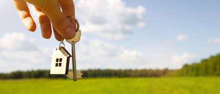 Wooden pendant of a house and key. Background of the sky and field. Dream of home, building a cottage in the countryside, plan and design, delivery of the project, moving to a new house. Copy spaceの写真素材