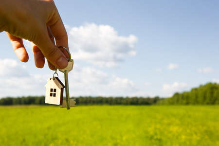 Wooden pendant of a house and key. Background of the sky and field. Dream of home, building a cottage in the countryside, plan and design, delivery of the project, moving to a new house. Copy spaceの写真素材