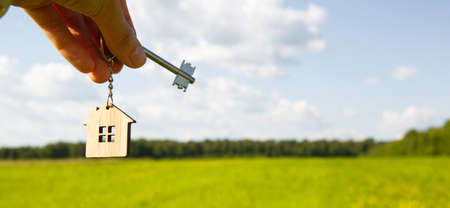 Wooden pendant of a house and key. Background of the sky and field. Dream of home, building a cottage in the countryside, plan and design, delivery of the project, moving to a new house. Copy spaceの写真素材