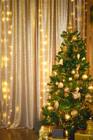 Christmas green artificial fir tree in the Golden room with lights garlands decorated with Golden balls, beads and toys. The box with the gifts in foil and black paper. New Year. Space for textの写真素材