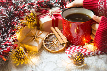 A woman's hand in a warm sweater holds a red mug with a hot drink on a table with Christmas decorations. New year's atmosphere, cinnamon sticks and a slice of dried orange, gifts, garland and tinselの写真素材