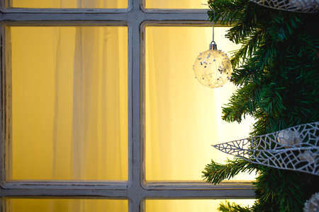 Christmas decorations made of spruce branches and white transparent balls and ribbons frame the window with wooden frame and yellow light. New year festive atmosphere, comfort of home. Space for textの写真素材