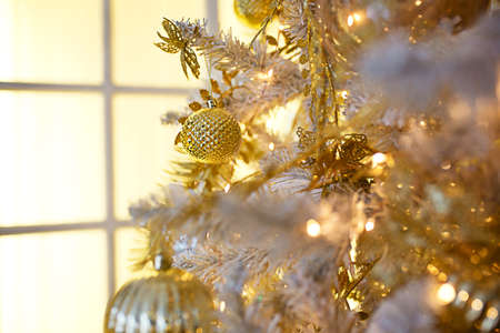 Gold Christmas decorations on a white artificial spruce close - up. Festive background, new year. Window with yellow light. Space for textの写真素材