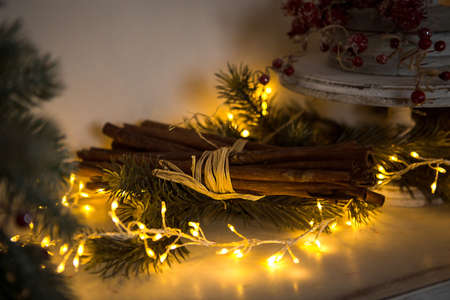 Cinnamon sticks tied with string in yellow lights garlands on Christmas tree branches and red berries. The atmosphere of the new year festive background, details of decorationsの写真素材