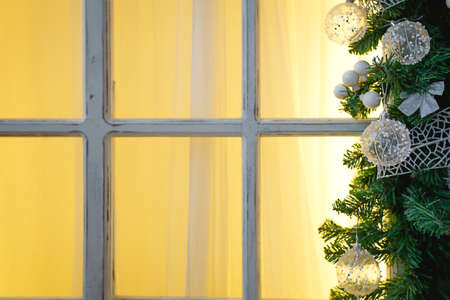 Christmas decorations made of spruce branches and white transparent balls and ribbons frame the window with wooden frame and yellow light. New year festive atmosphere, comfort of home. Space for textの写真素材