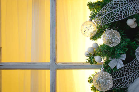Christmas decorations made of spruce branches and white transparent balls and ribbons frame the window with wooden frame and yellow light. New year festive atmosphere, comfort of home. Space for textの写真素材