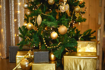 Christmas green artificial fir tree in the Golden room with lights garlands decorated with Golden balls, beads and toys. The box with the gifts in foil and black paper. New Year. Space for textの写真素材