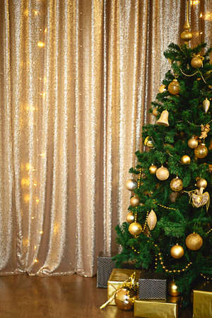Christmas green artificial fir tree in the Golden room with lights garlands decorated with Golden balls, beads and toys. The box with the gifts in foil and black paper. New Year. Space for textの写真素材