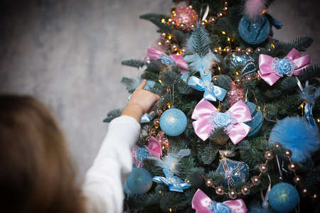 A little girl reaches for a Christmas tree toy with her hand. Christmas decor, waiting for a holiday and a miracle. New Year. White, pink and blue decor, Golden lights garlandsの写真素材
