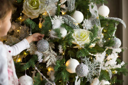 A little girl reaches for a Christmas tree toy with her hand. Christmas decor, waiting for a holiday and a miracle. New Year. White, pink and blue decor, Golden lights garlandsの写真素材