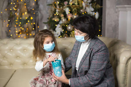 Grandmother and granddaughter on the sofa in the living room with Christmas decor hugging in medical masks on their faces. A family holiday during the outbreak of coronavirus and disease. New Yearの写真素材