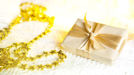 Box with Christmas gift in light paper is tied with a gold ribbon on white knitted background with decor. Golden garland of stars. New Year. Congratulations, surprise. space for textの写真素材