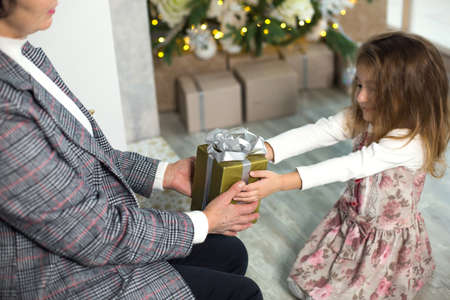 Grandma gives her granddaughter a gift for Christmas. The girl stretches out her hands to the box with the bow. New year, family holiday, communication of generationsの写真素材